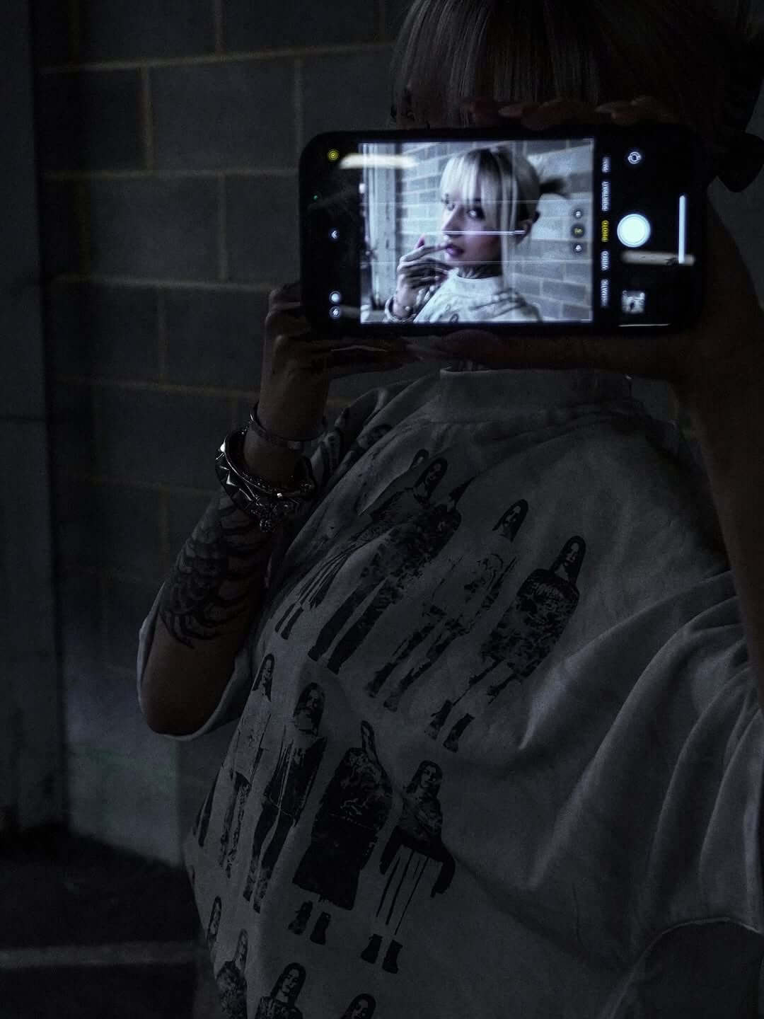 Person holding a phone to capture a selfie, dressed in a graphic tee with a vintage design.