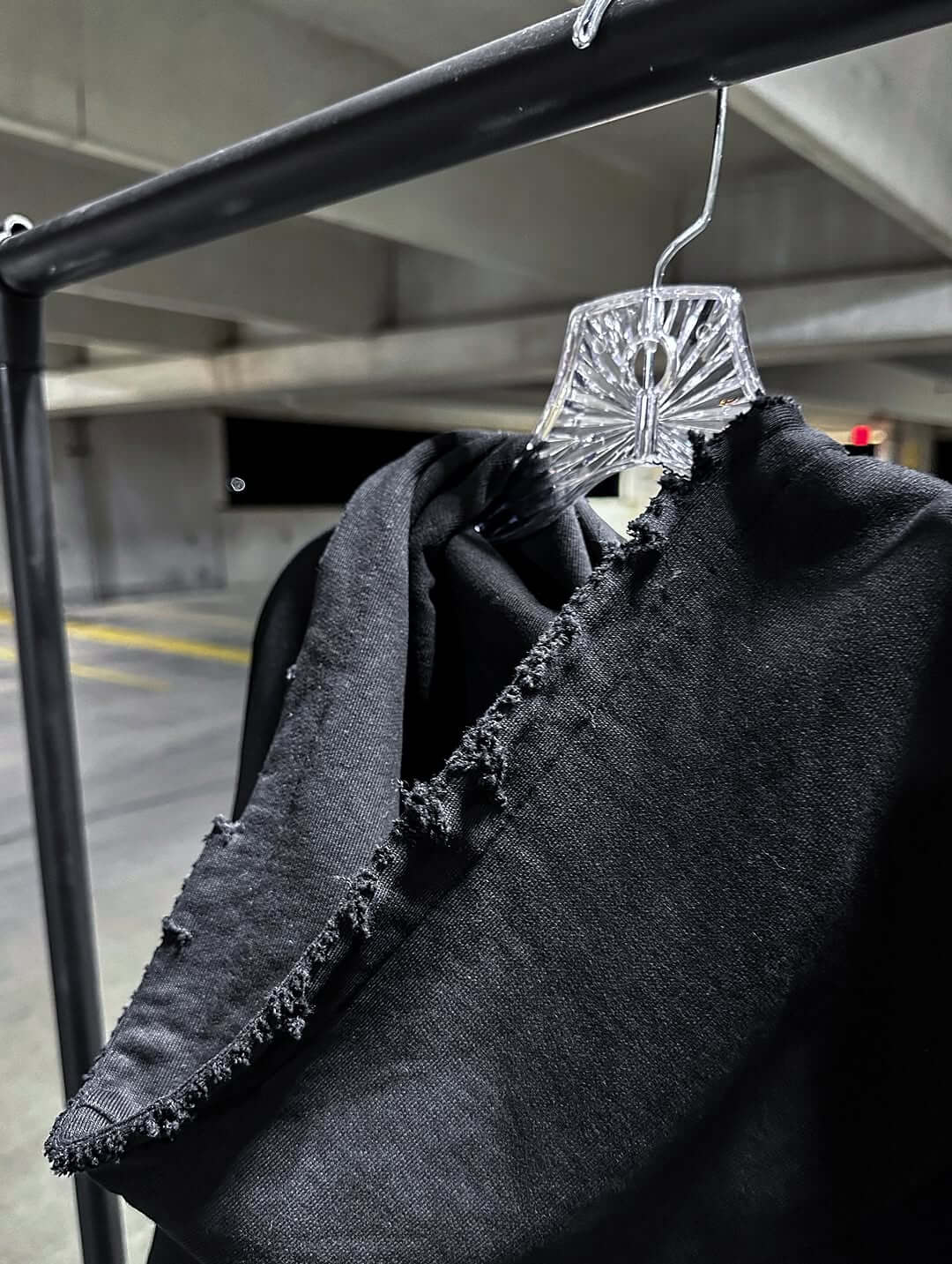 Black oversized hoodie showcasing raw cut hem detail, hanging on a clear plastic hanger in a parking structure.